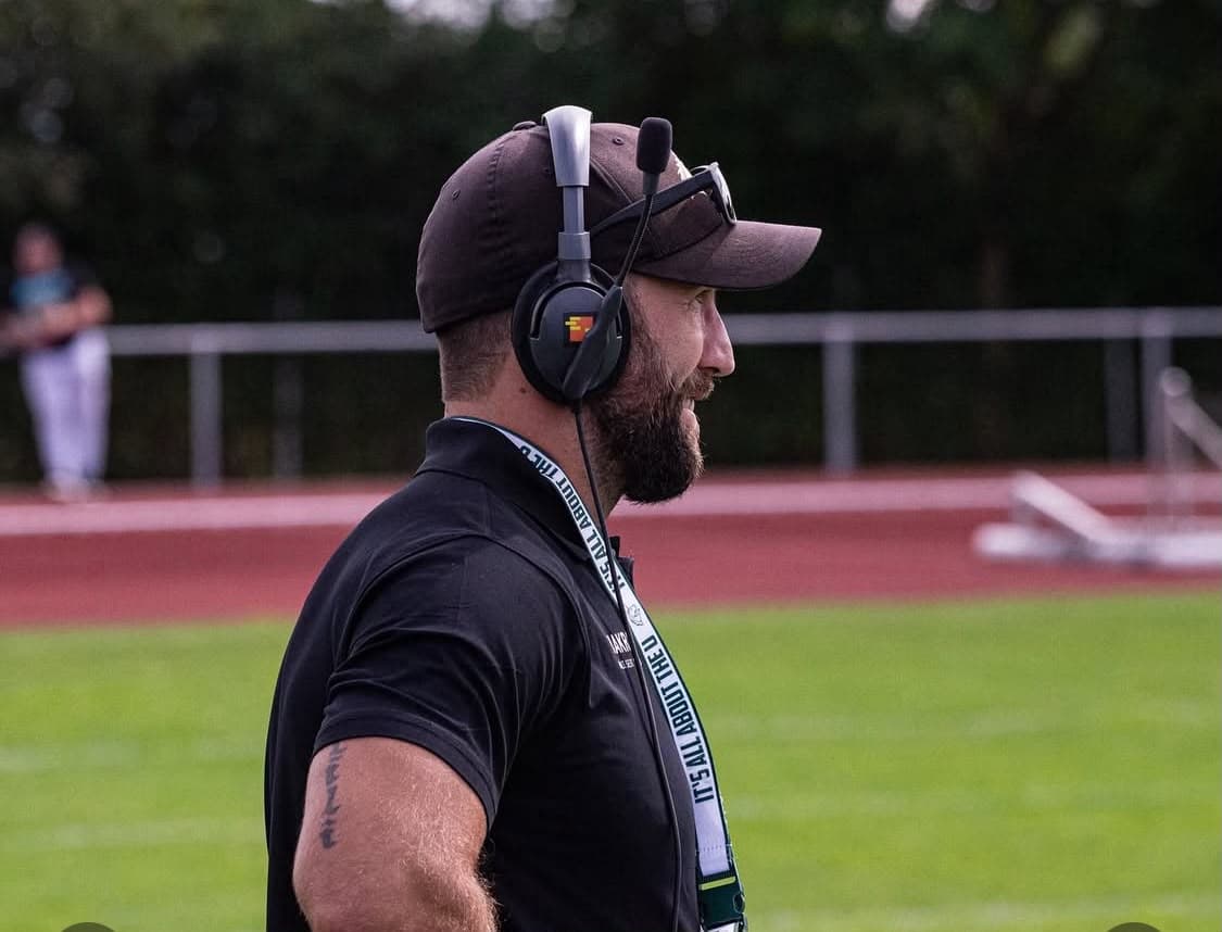 Michael Gentilli in profile on the sideline wearing a coaching headset